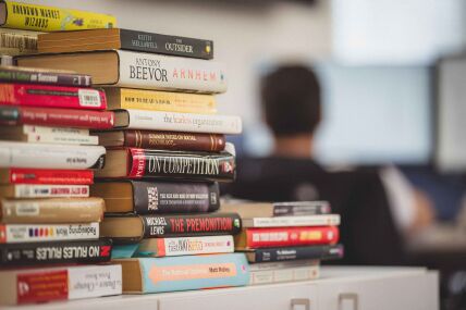 Three uneven stacks of books of multiple sizes and colours rests on a table, with a person seated in front out of focus looking at a computer monitor.