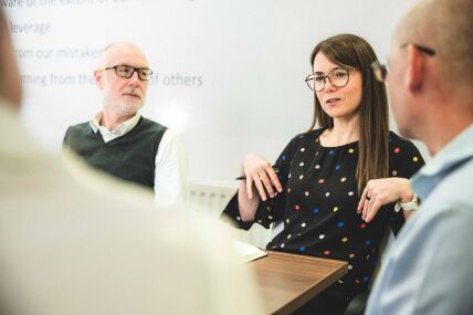 Lady with glasses and long brown hair, wearing a black polka dot top and holding a pen, sat at a table next to a man with glasses and a grey beard