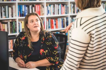 A woman with a black floral shirt and blonde shoulder length hair engages in conversation with another woman in a white and black striped top while seated at a desk with a bookcase full of books behind